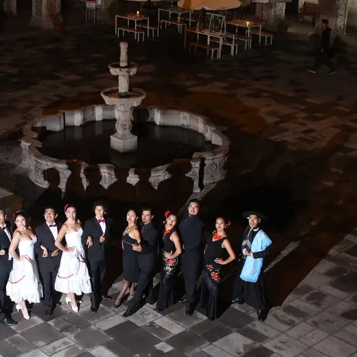 Elenco de marinera con vestidos blancos y trajes oscuros alrededor de una pileta en un patio de noche