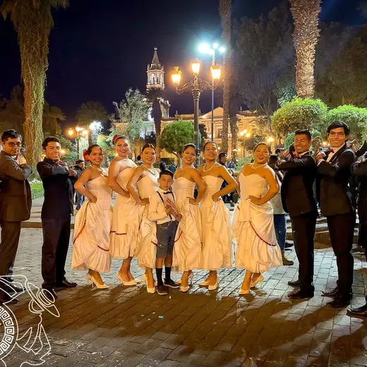Elenco con vestidos claros y trajes oscuros posando de noche en una plaza
