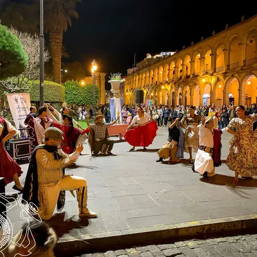 Presentación de danza en una plaza de noche, con público y arcos iluminados