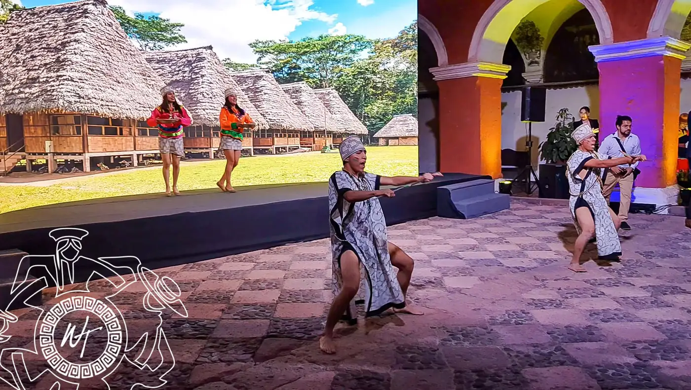 Bailarines con vestuario amazónico y andino sobre un escenario con casas de paja de fondo, en un claustro colonial